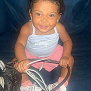 Zarahi is registered to the contest to win money with this photo: child, toddler, baby, smiling, portrait, sitting, indoors, couch, dark_background, curly_hair, hair_buns, pink_clothing, tank_top, skirt, sneakers, socks, handbag, cables, flash_photography, happy