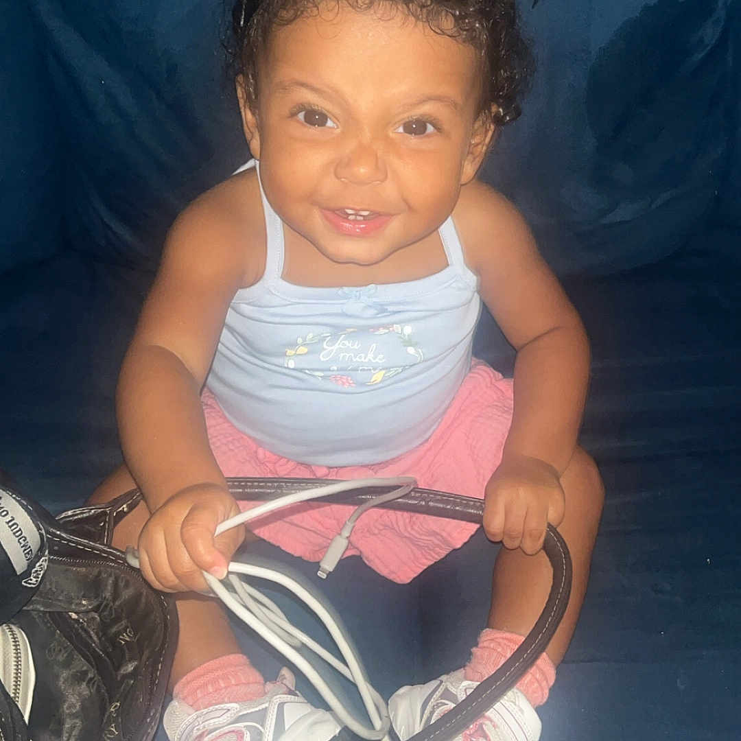 Zarahi is registered to the contest to win money with this photo: child, toddler, baby, smiling, portrait, sitting, indoors, couch, dark_background, curly_hair, hair_buns, pink_clothing, tank_top, skirt, sneakers, socks, handbag, cables, flash_photography, happy