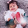 baby, smiling, pink_bow, hair_accessory, white_outfit, red_design, colorful_shoes, fluffy_cushion, gray_cushion, child, indoor, portrait, happy, cute, infant, sitting, soft_texture, cozy, person, young_child
