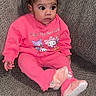 baby, child, pink_clothing, hello_kitty, sweatshirt, pants, hair_buns, pink_bows, couch, textured_fabric, footwear, fur_booties, indoor, person, seated, curious_expression, hand, small_child, portrait, casual_wear