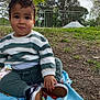 baby, blanket, casual_clothing, child, curious, daylight, fence, grass, green_pants, ground, nature, outdoor, park, person, shoes, sitting, striped_sweater, toddler, trees, young_child