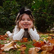Mélina a rejoint le concours — aidez-le/la à gagner de superbes lots ! autumn, child, face, fall, girl, grass, greenery, hair_bow, hands, happy, leaves, lying_down, nature, outdoor, playful, portrait, seasonal, smile, white_sweater, young