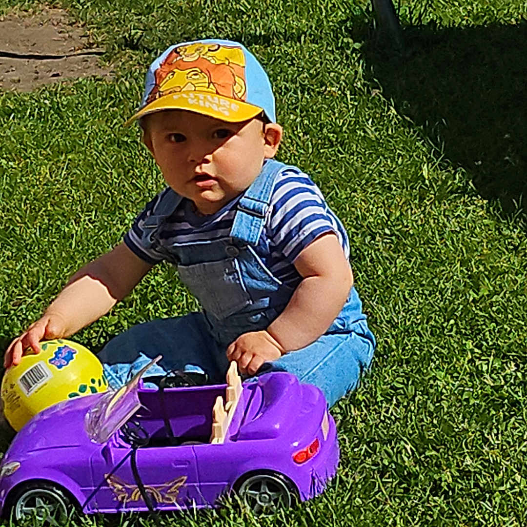 Loukas a rejoint le concours — aidez-le/la à gagner de superbes lots ! backyard, ball, cap, casual_clothing, child, daylight, fence, fun, garden, grass, greenery, outdoor, overalls, play, sitting, summer, sunlight, toddler, toy_car, young_child