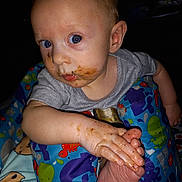 Connor is registered to the contest to win money with this photo: baby, blue_eyes, child, close_up, colorful_baby_seat, curious_expression, food_smudges, foot, gray_shirt, hand, indoor, infant, messy_face, person, playful, portrait, sea_creatures, sitting, skin, young_child