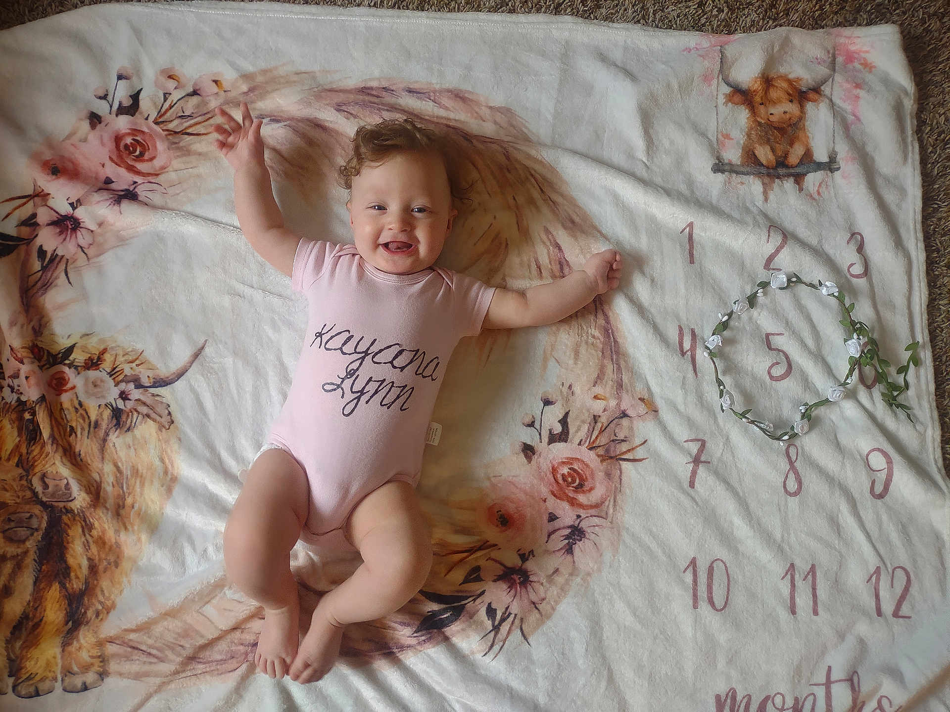 Kayana joined the competition — help win amazing prizes! baby, blanket, carpet, cheerful, cute, floral, hair, hand_raised, highland_cow, illustration, infant, lying_down, milestone_blanket, numbers, onesie, pink, portrait, smiling, toes, wreath