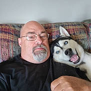 Thor is registered to the contest to win money with this photo: man, dog, husky, blue_eyes, glasses, bald, beard, couch, indoor, pet, portrait, relaxing, black_shirt, happy, face, smiling_dog, furniture, closeup, friendship, home