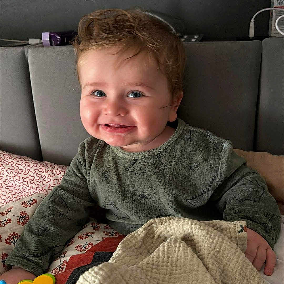 Noah a rejoint le concours — aidez-le/la à gagner de superbes lots ! baby, child, smiling, curly_hair, blanket, toy, bed, green_shirt, indoors, happy, cute, person, toddler, face, hand, cozy, comfort, playful, portrait, young