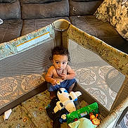 Julian joined the competition — help win amazing prizes! toddler, child, playpen, toy, airplane_toy, stuffed_animal, couch, water_bottle, snack, curly_hair, indoor, carpet, blanket, netting, blue_pants, necklace, seated, looking_up, home, floor