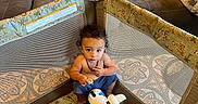 Julian joined the competition — help win amazing prizes! airplane_toy, blanket, blue_pants, carpet, child, couch, curly_hair, floor, home, indoor, looking_up, necklace, netting, playpen, seated, snack, stuffed_animal, toddler, toy, water_bottle