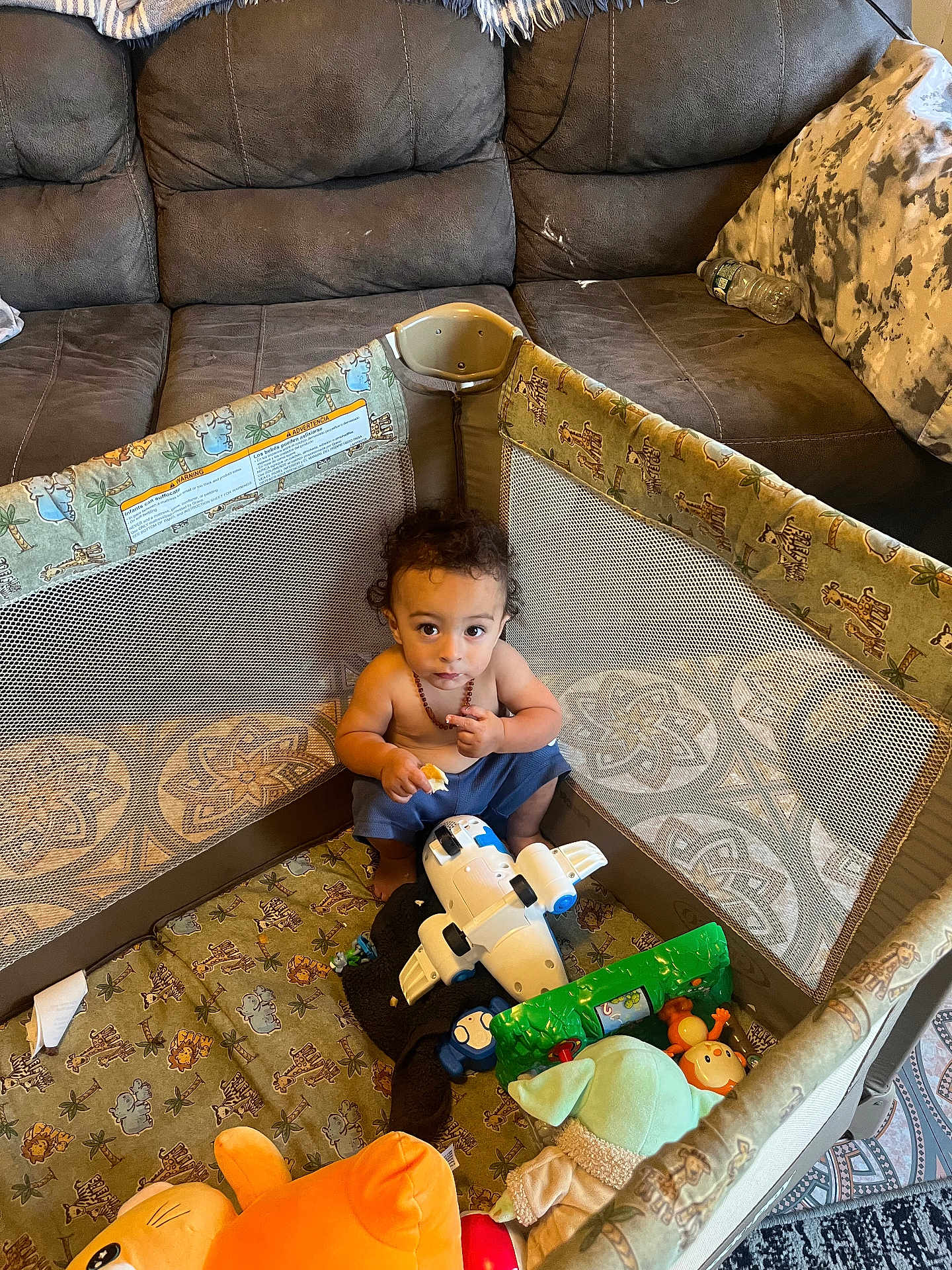 Julian joined the competition — help win amazing prizes! toddler, child, playpen, toy, airplane_toy, stuffed_animal, couch, water_bottle, snack, curly_hair, indoor, carpet, blanket, netting, blue_pants, necklace, seated, looking_up, home, floor
