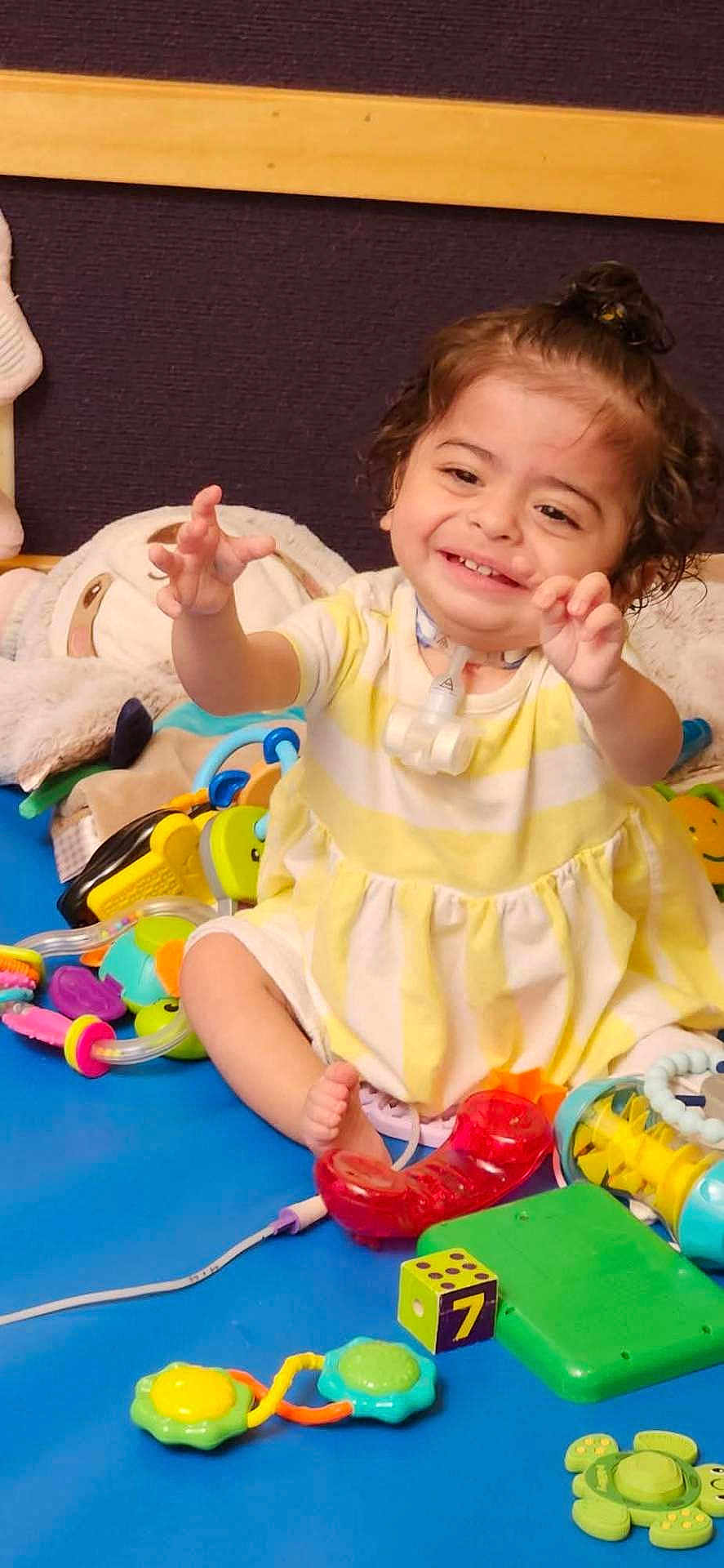 Athena joined the competition — help win amazing prizes! child, toddler, smiling, curly_hair, yellow_dress, sitting, toys, colorful, indoor, happy, playtime, baby_toys, floor_mat, cute, infant, reaching_out, person, young_child, joyful, play