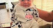 Levi is registered to the contest to win money with this photo: baby, infant, cow_print, onesie, table, wood, curtains, window, sunlight, person, arm, milk_bottle, phone, indoor, child, cute, expression, sitting, clothing, home