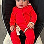baby, infant, red_onesie, headband, strawberry_pattern, baby_rocker, buckle, indoor, floor, slippers, curious, seated, clothing, child, person, cute, portrait, hands, footwear, carpet