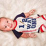 baby, infant, smiling, lying_down, blanket, clothing, baseball_theme, onesie, cute, child, portrait, happy, soft_texture, baby_garment, person, small_hands, head, face, eyes, mouth