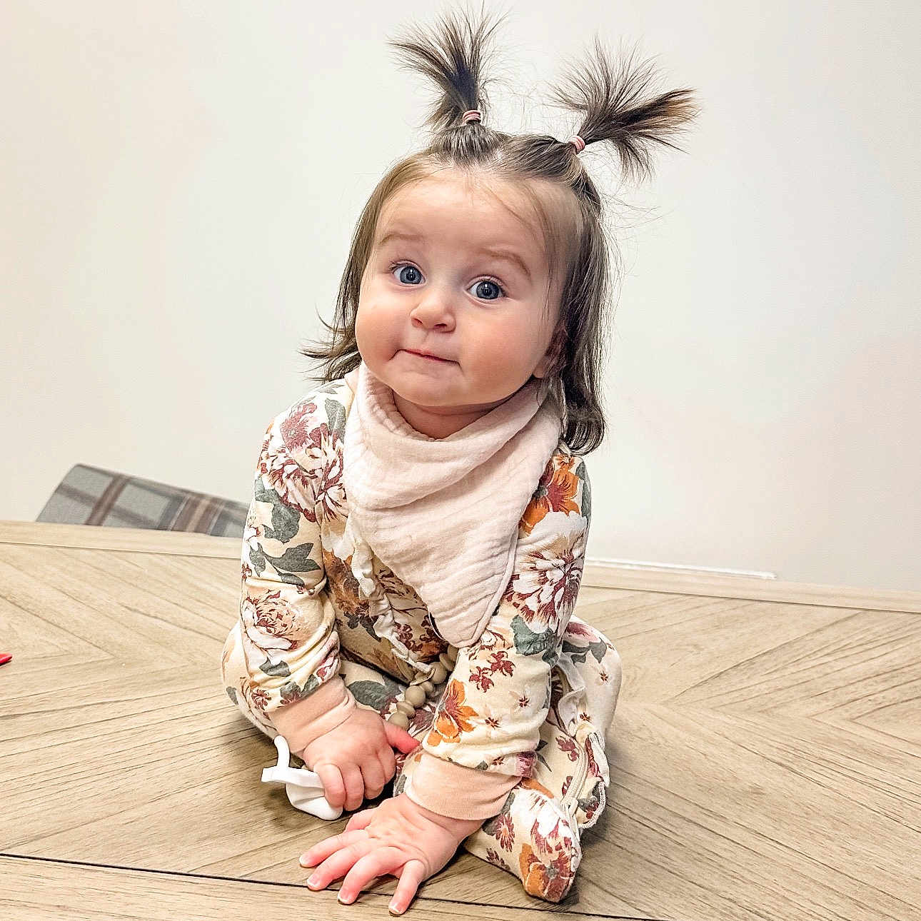 Monroe is registered to the contest to win money with this photo: baby, bib, big_eyes, child, chubby_cheeks, clothing, cute, floral_onesie, hair_ties, hands, indoor, neutral_background, pacifier, pigtails, playful, portrait, sitting, soft_lighting, toddler, wooden_table
