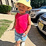 toddler, child, yellow_hat, red_shirt, blue_shorts, white_shoes, outdoor, sidewalk, sunny, daytime, parked_cars, greenery, smiling, walking, person, casual_clothing, hat, cheerful, holding_object, urban