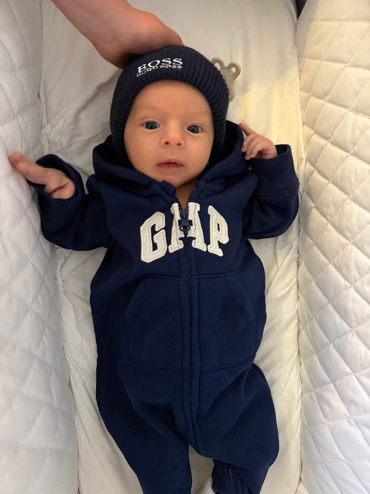 Jack is registered to the contest to win money with this photo: baby, infant, onesie, navy_blue, beanie, hat, clothing, face, head, person, quilted, white, bed, hand, newborn, sleeping_area, cute, portrait, babywear, soft