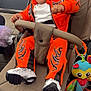 baby, stroller, orange_hoodie, orange_pants, white_shoes, laughing, happy, child, toy, plush_toy, outdoor, pavement, seatbelt, casual_clothing, person, infant, smile, curly_hair, footwear, daylight