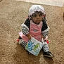 child, baby, carpet, book, reading, indoor, floor, mop, bonnet, striped_clothing, pink_dress, black_shoes, curious, serious, seated, home, toy, flooring, young, portrait