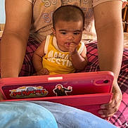Waziya is registered to the contest to win money with this photo: baby, child, tablet, adult, hands, yellow_shirt, plaid_pants, indoor, curious, technology, face, person, sitting, holding, arm, close_up, home, furniture, skin, expression