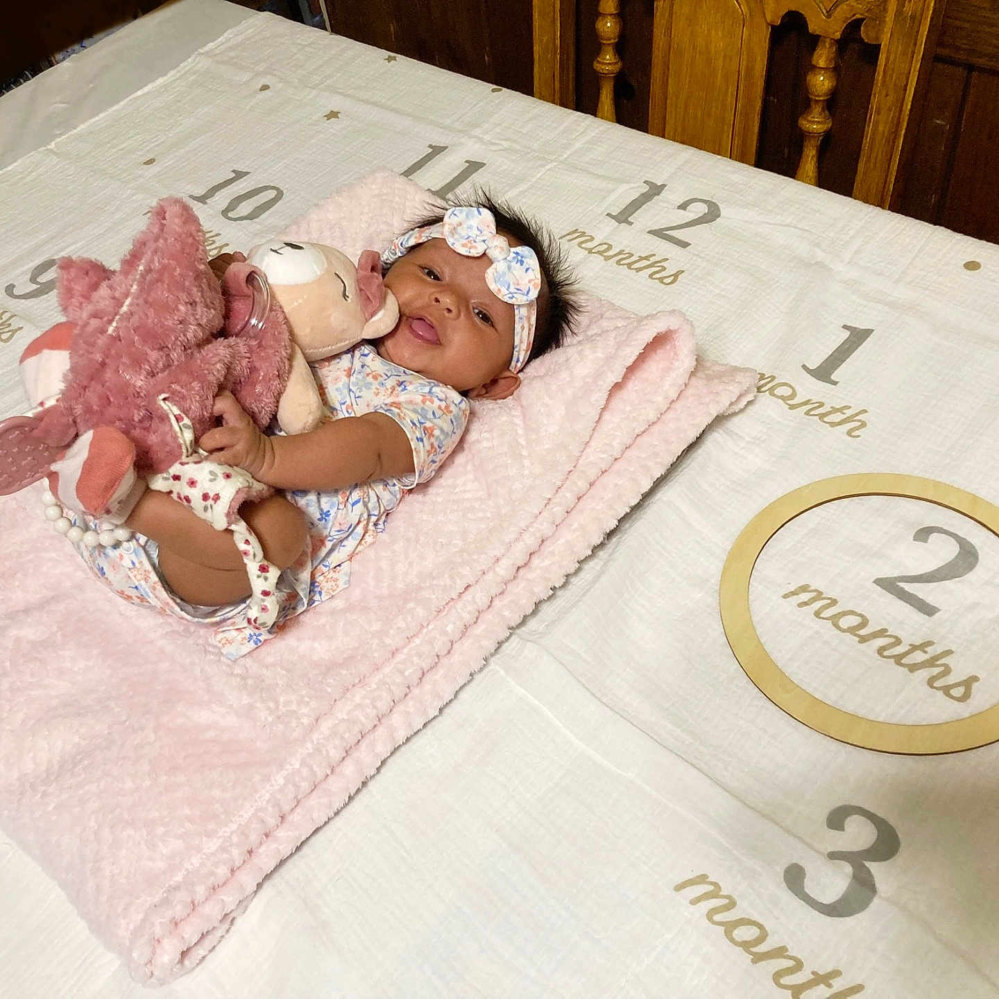 Rayla joined the competition — help win amazing prizes! baby, infant, blanket, plush_toy, headband, clothing, milestone_cloth, wooden_ring, pink, soft_texture, smiling, lying_down, indoors, wooden_chair, table, face, person, cute, child, decor