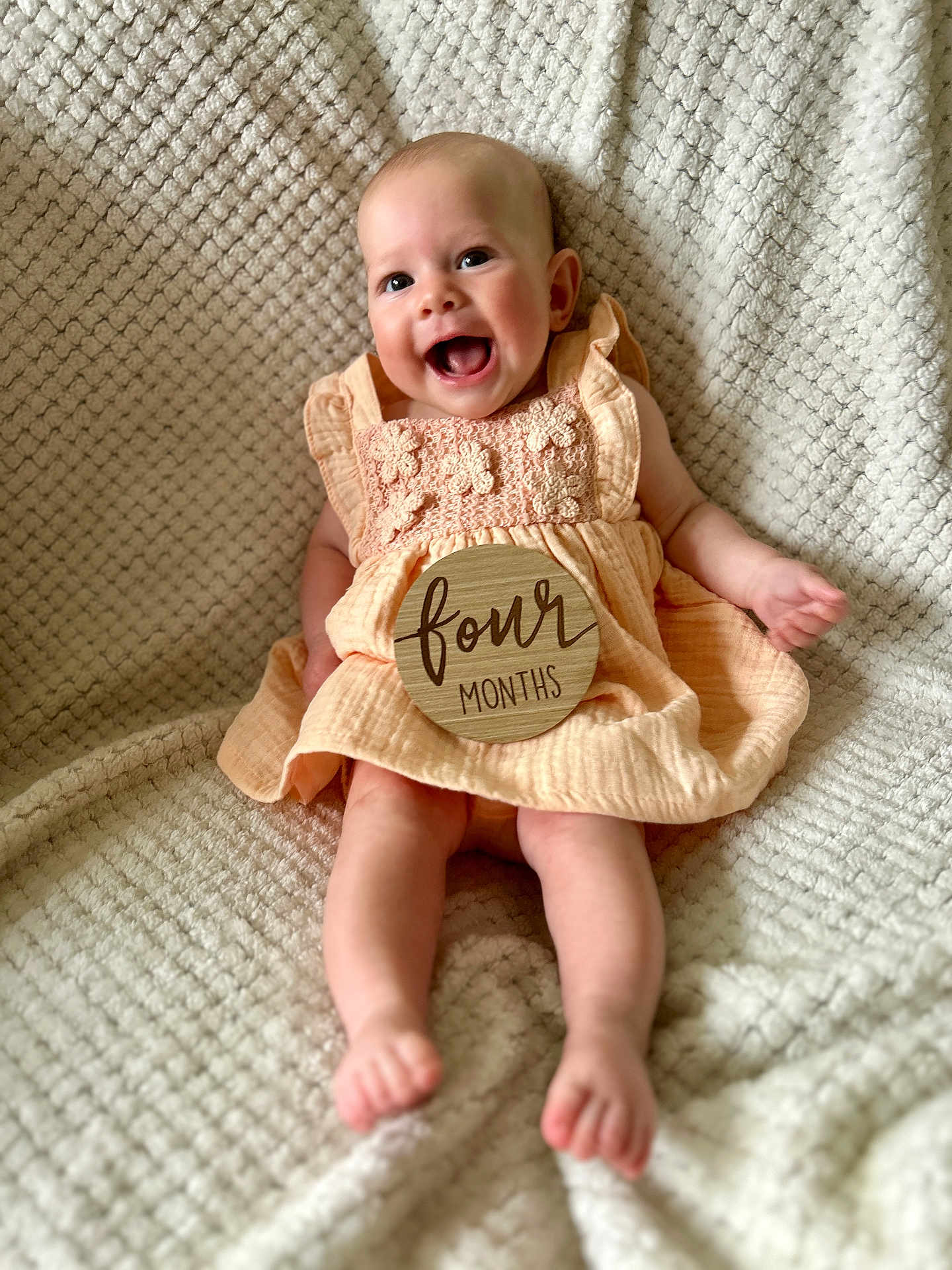 Madelyn is registered to the contest to win money with this photo: baby, infant, smile, dress, peach, blanket, texture, wooden_sign, four_months, happy, cute, sitting, child, indoors, portrait, face, skin, feet, hand, young