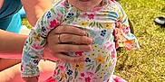 Madelyn is registered to the contest to win money with this photo: baby, child, person, floral_swimsuit, sunhat, blanket, hand, grass, lake, water, outdoor, toy, pink, summer, nature, cute, expression, holding, sitting, family