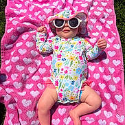 Madelyn is registered to the contest to win money with this photo: baby, towel, pink, hearts, sunglasses, hat, floral, swimsuit, outdoor, grass, sunny, child, relaxing, cute, summer, babywear, barefoot, infant, lying_down, colorful