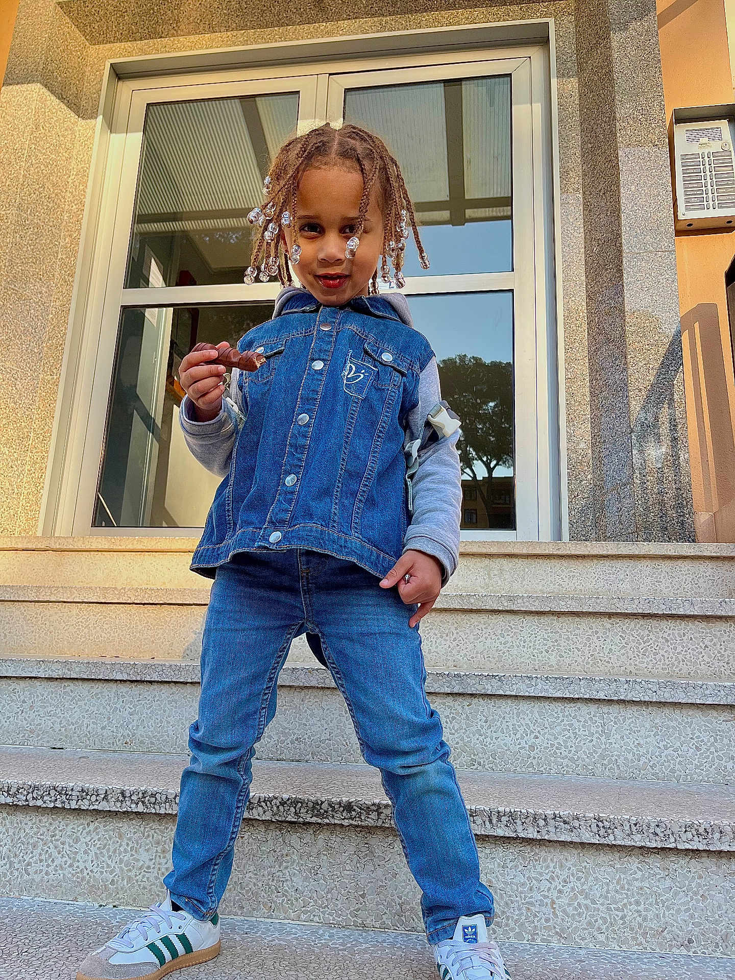 Noah a rejoint le concours — aidez-le/la à gagner de superbes lots ! child, denim_vest, jeans, sneakers, stairs, building, chocolate_bar, braided_hair, beads, smiling, outdoor, casual_clothing, person, fashion, playful, sunlight, modern_architecture, steps, young_child, portrait