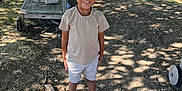 Noah participe au concours pour gagner de l'argent avec cette photo : bag, bench, casual_clothing, child, daytime, dirt, grass, hairstyle, happy, nature, outdoor, park, person, shoes, shorts, smile, standing, sunlight, tree_shadow, tshirt