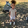 child, outdoor, sunlight, tree_shadow, bench, bag, grass, dirt, smile, shorts, tshirt, shoes, hairstyle, person, daytime, nature, park, standing, casual_clothing, happy