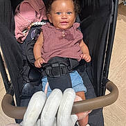 Maïna a rejoint le concours — aidez-le/la à gagner de superbes lots ! baby, stroller, child, smiling, curly_hair, denim_shorts, mauve_top, sandal, toy, spiral_toy, black_harness, indoor, wooden_floor, accessory, sunglasses, pink_cloth, face, person, happy, seated