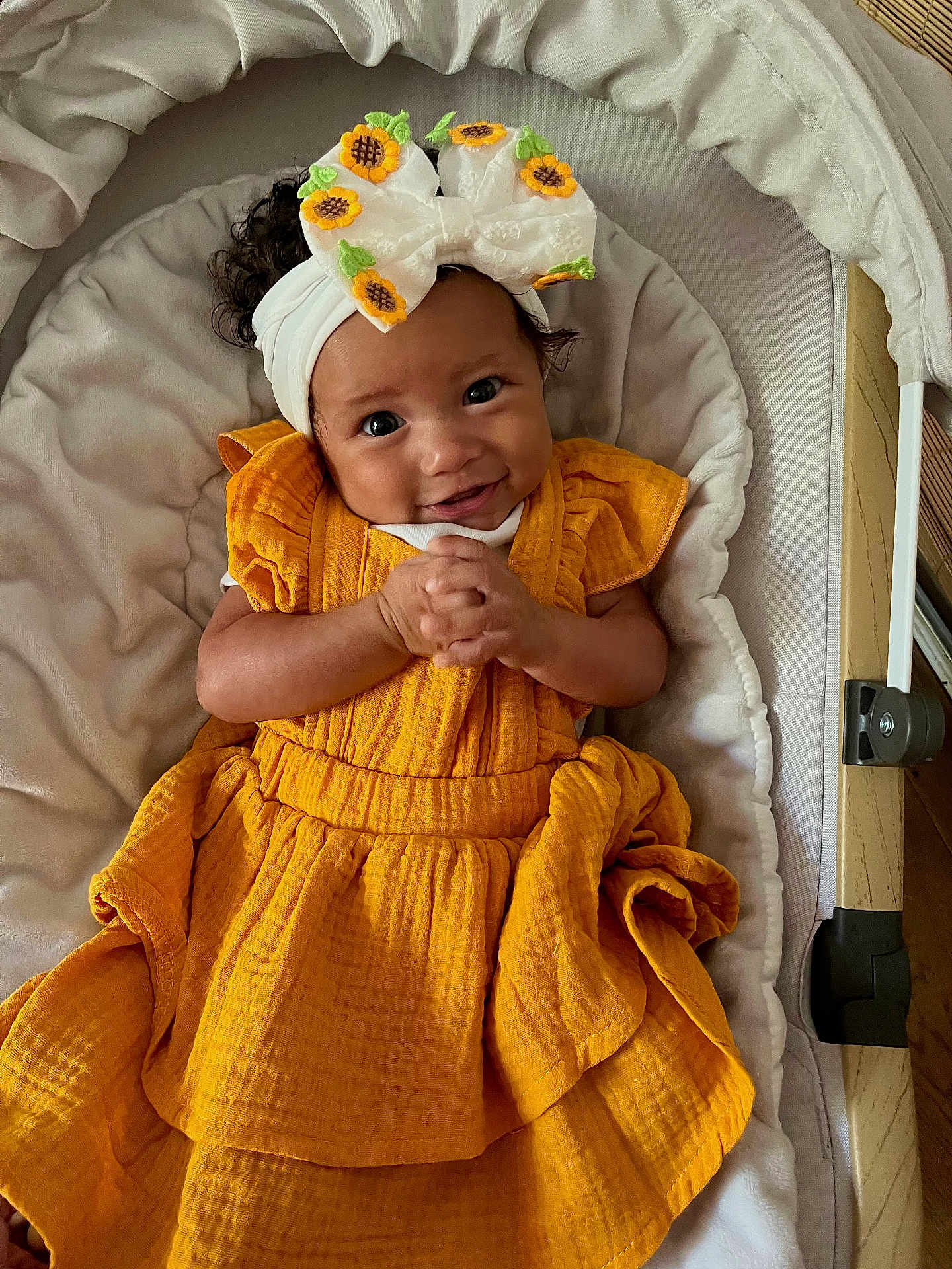 Maïna a rejoint le concours — aidez-le/la à gagner de superbes lots ! baby, infant, yellow_dress, headband, sunflower, smiling, hands_clasped, cushion, baby_seat, curly_hair, comfortable, indoor, portrait, cute, happy, child, person, fashion, apparel, closeup