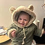 baby, child, smiling, coat, hood, fur_trim, bear_ears, indoor, person, clothing, cute, warm, seated, expression, portrait, hand, face, furniture, floor, home