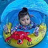 baby, blue, child, colorful, crab, curious, cute, floatation_device, floral_pattern, infant, inflatable_float, outdoor, playful, pool, sitting, summer, swimsuit, topknot, toy, water