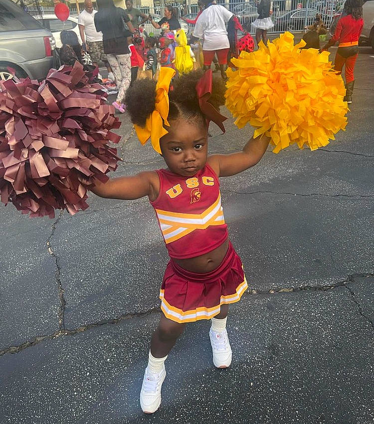Harlee is registered to the contest to win money with this photo: asphalt, child, costume, dance, event, festival, flower, fun, human_leg, pedestrian, person, public_event, road, sandal, shorts, sock, street, thigh, toddler, wheel