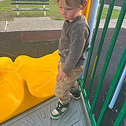 Tate is registered to the contest to win money with this photo: bench, casual_clothing, child, concrete, daylight, grass, green_railings, outdoor, pants, park, person, play_equipment, playground, sidewalk, slide, sneakers, sweater, thoughtful, yellow, young_boy