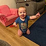 baby, child, infant, smiling, happy, kneeling, indoor, living_room, portrait, face, blue_shirt, onesie, toy, armchair, sofa, car_seat, rug, wooden_floor, home, sitting