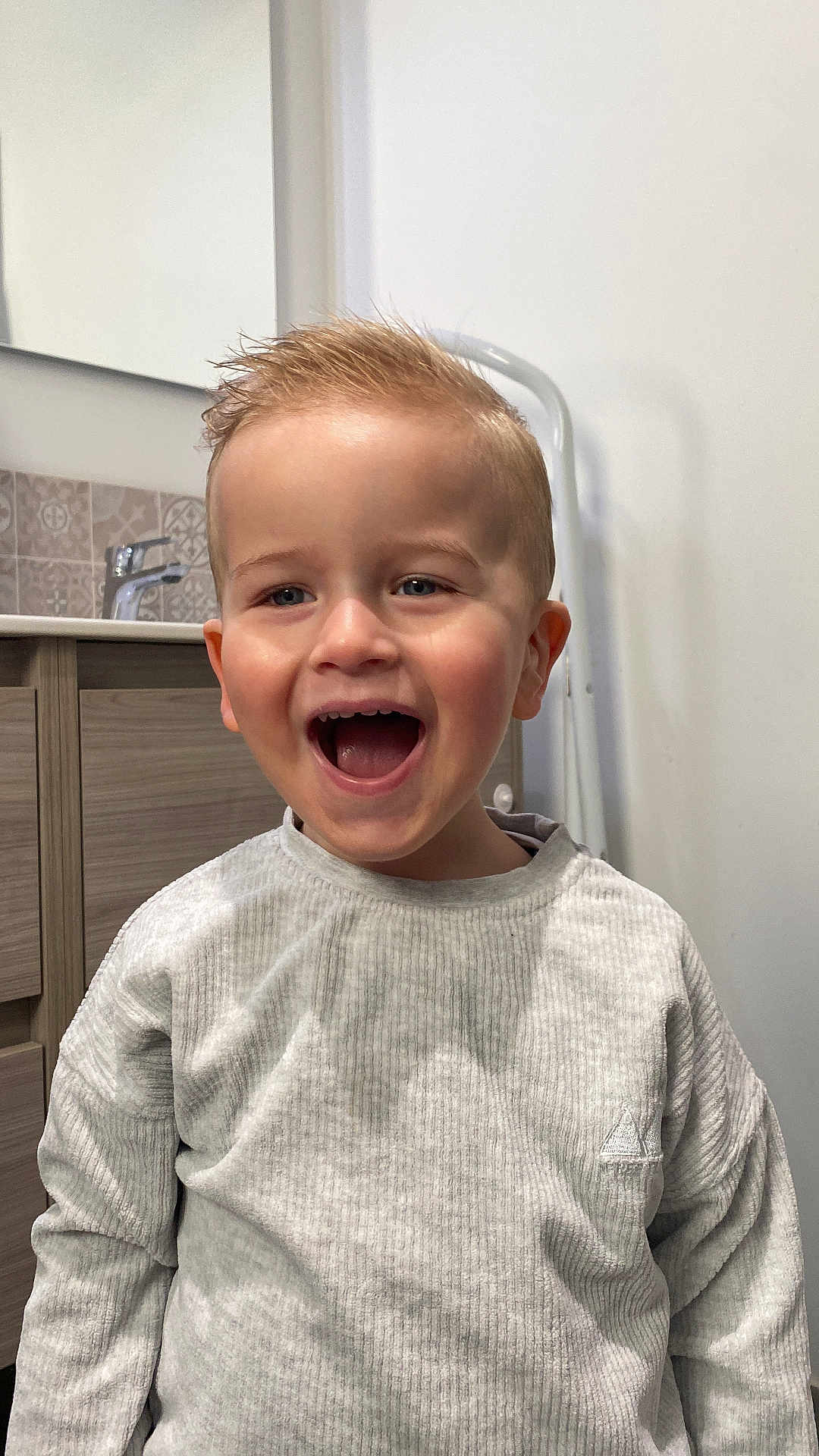 Aykan participe au concours pour gagner de l'argent avec cette photo : toddler, child, smiling, happy, indoor, bathroom, sink, mirror, light_grey_sweater, short_hair, blonde_hair, open_mouth, person, face, joyful, casual_clothing, modern, tile, cabinet, home