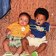 António is registered to the contest to win money with this photo: toddler, baby, child, couch, orange_shirt, blue_shirt, pacifier, plaid_shorts, white_shorts, green_shoes, indoor, seated, curly_hair, short_hair, looking_away, looking_at_camera, furniture, smiling, cute, relaxed