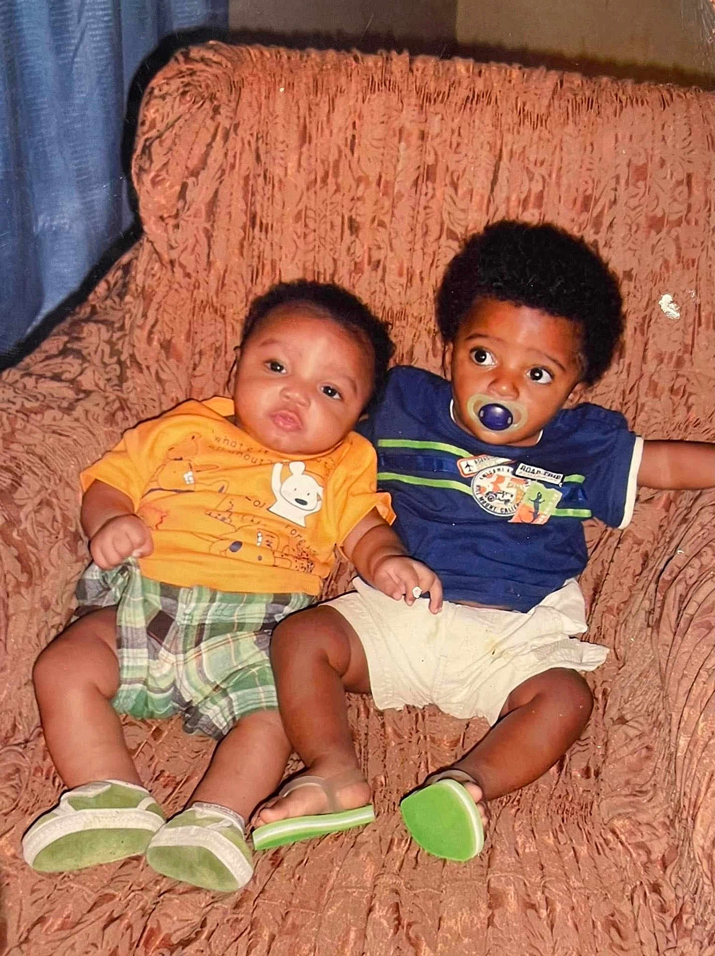 António is registered to the contest to win money with this photo: toddler, baby, child, couch, orange_shirt, blue_shirt, pacifier, plaid_shorts, white_shorts, green_shoes, indoor, seated, curly_hair, short_hair, looking_away, looking_at_camera, furniture, smiling, cute, relaxed
