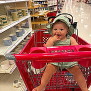 Chanel Leigh joined the competition — help win amazing prizes! baby, shopping_cart, store, aisle, mugs, dishes, green_hat, green_outfit, smiling, child, indoor, retail, floor, sitting, footwear, hand, finger_in_mouth, red_cart, person, happy