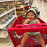 baby, shopping_cart, store, aisle, mugs, dishes, green_hat, green_outfit, smiling, child, indoor, retail, floor, sitting, footwear, hand, finger_in_mouth, red_cart, person, happy