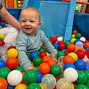 Dominic Nicolas is registered to the contest to win money with this photo: toddler, child, ball_pit, colorful_balls, blue_sweater, smiling, happy, indoor_play_area, toy, play, fun, person, arm, white_pants, orange_balls, green_balls, blue_balls, yellow_balls, red_balls, plastic_balls