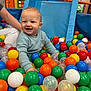 toddler, child, ball_pit, colorful_balls, blue_sweater, smiling, happy, indoor_play_area, toy, play, fun, person, arm, white_pants, orange_balls, green_balls, blue_balls, yellow_balls, red_balls, plastic_balls