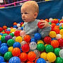 toddler, child, ball_pit, colorful_balls, play_area, indoor, cute, curious, young_child, plastic_balls, blue_sweater, red_ball, yellow_ball, green_ball, orange_ball, smiling, face, fun, leisure, recreation