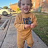 toddler, child, smiling, curly_hair, sidewalk, outdoor, sneakers, brown_clothing, building, brick_wall, grass, daylight, happy, person, casual_wear, blue_sky, parked_car, urban, cute, walking