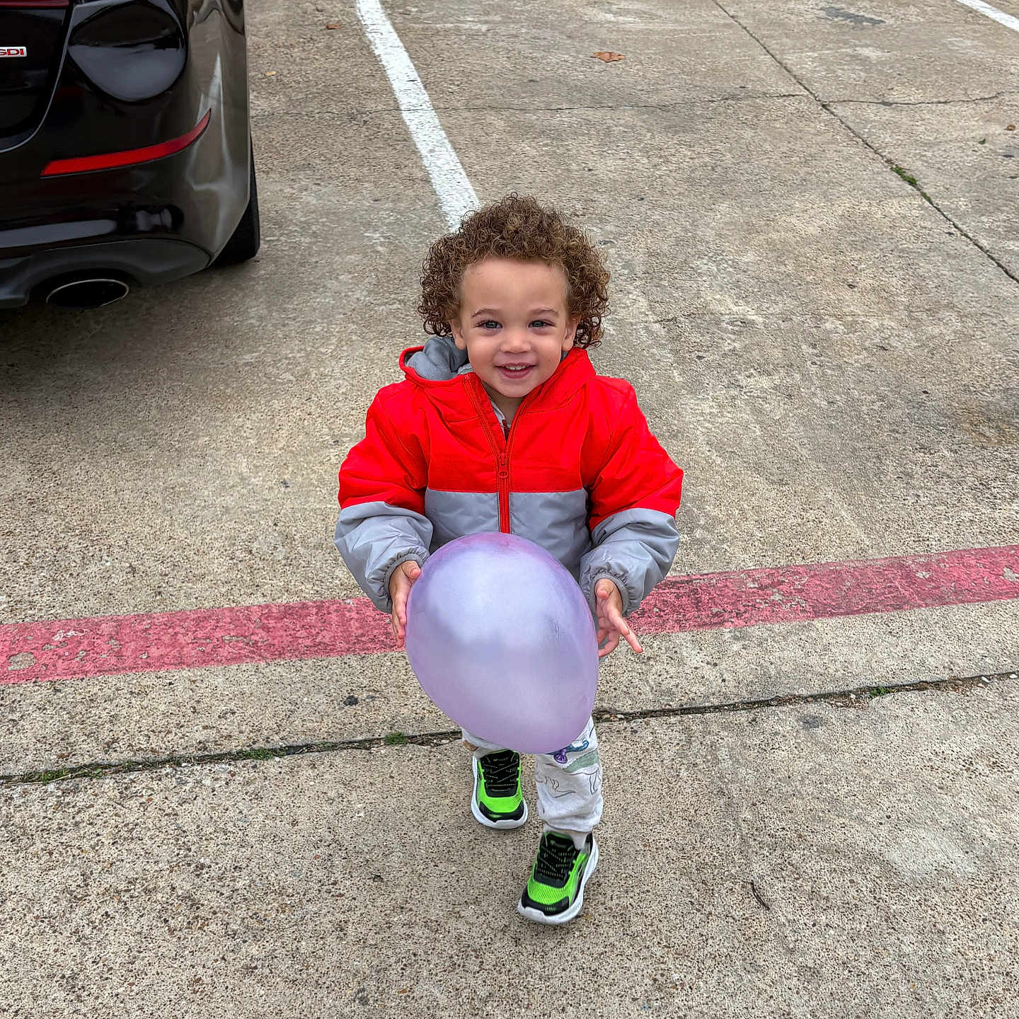 Adam is registered to the contest to win money with this photo: child, toddler, balloon, curly_hair, red_jacket, gray_jacket, sneakers, parking_lot, car, concrete, outdoor, smiling, holding, playful, young_child, sidewalk, street, vehicle, casual_clothing, daylight