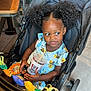 Adhara is registered to the contest to win money with this photo: toddler, child, stroller, curly_hair, drink_cup, toy, indoor, floor_tile, clothing, table, chair, person, shoes, happy, cute, seated, beverage, plastic_cup, fast_food, restaurant