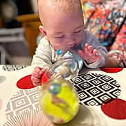 Djuliane a rejoint le concours — aidez-le/la à gagner de superbes lots ! adult, baby, blurred_background, bottle, child, clothing, colorful, curious, exploration, face, focus, hand, head, indoor, pattern, person, playing, skin, tablecloth, toy